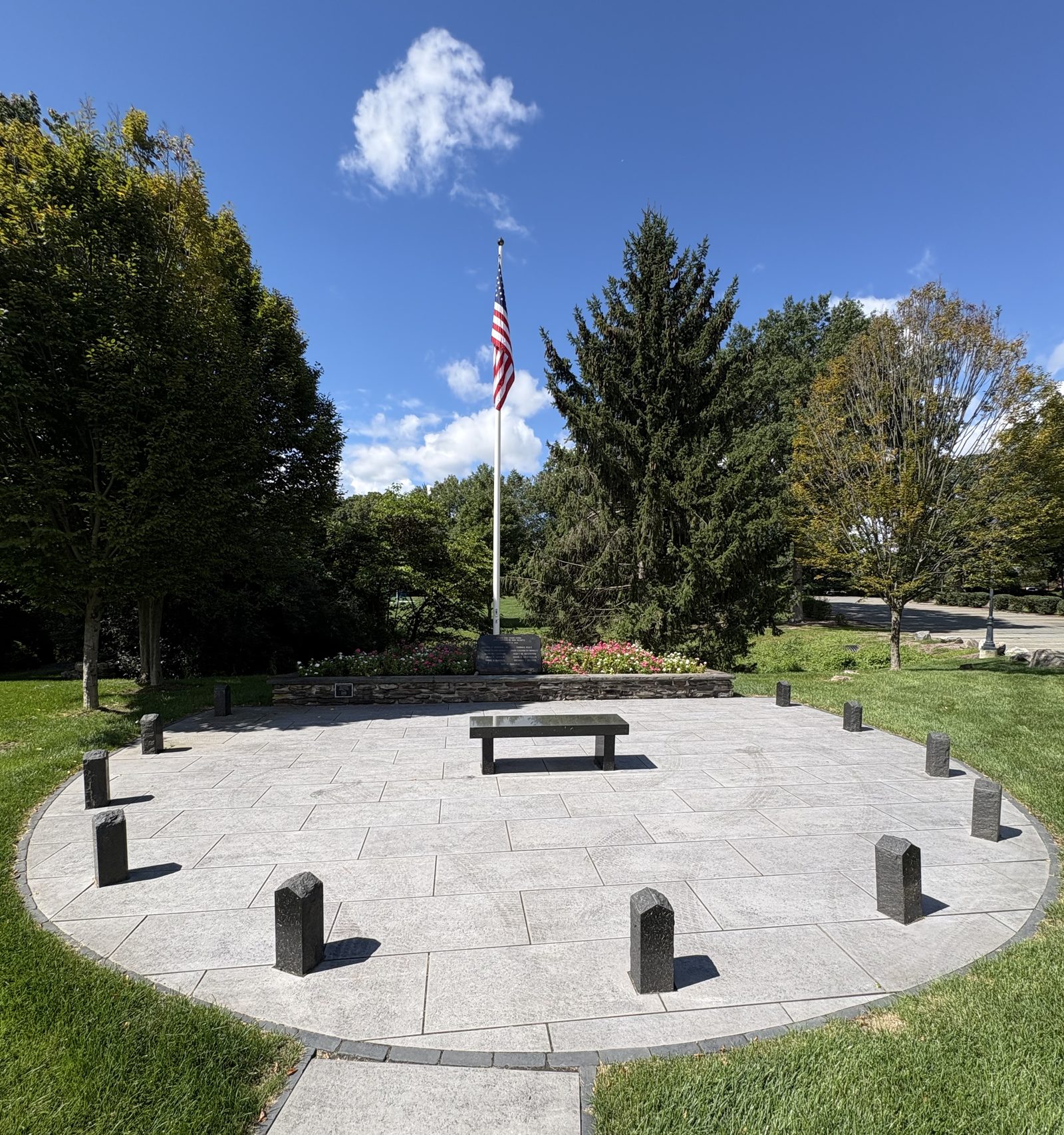 An oval paved plaza in Wyckoff Community Park ringed by eleven dark granite posts, with a low granite bench at its center and an American flag flying from a flagpole behind a fieldstone wall planted with flowers.