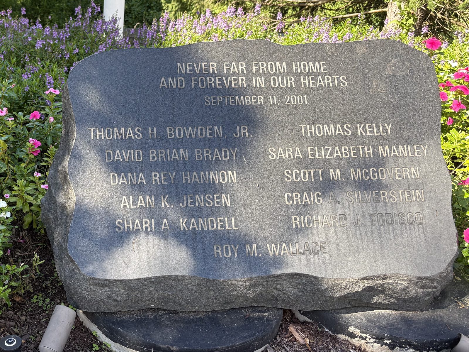 An inscribed granite stone reading “Never Far From Home and Forever in Our Hearts, September 11, 2001,” above the names of eleven people, set in a bed of pink and white flowers.