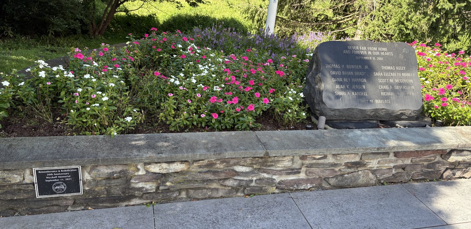 The memorial stone at the right end of a fieldstone wall planted with pink and white flowers, with a small dark plaque set into the wall at the left.